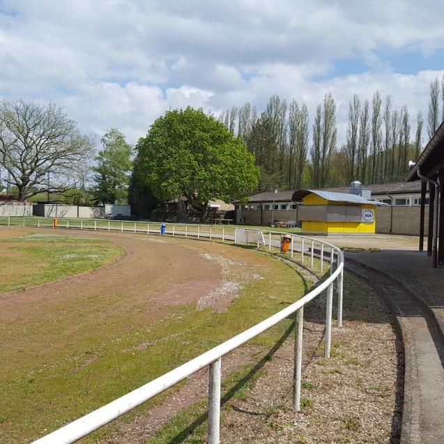 Blick auf einen Sportplatz mit Pferderennbahn, B&auml;umen und Nebengeb&auml;uden, umgeben von einer wei&szlig;en Umfriedung.