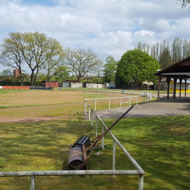 Blick auf einen Fu&szlig;ballplatz mit Toren, einem Weg, B&auml;umen und einem &uuml;berdachten Sitzbereich im Hintergrund.