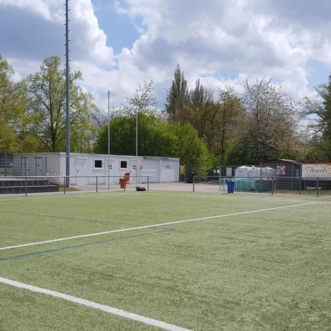 Leerstehender Sportplatz mit Kunstrasen, B&auml;nken, Geb&auml;uden im Hintergrund und gr&uuml;nem Baumbestand.