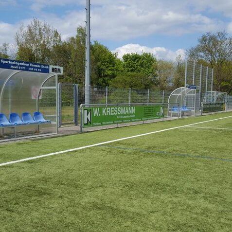 Blick auf eine Fu&szlig;ballbank mit blauen Sitzen und Werbebanner auf einem Kunstrasenplatz unterhalb eines blauen Himmels.