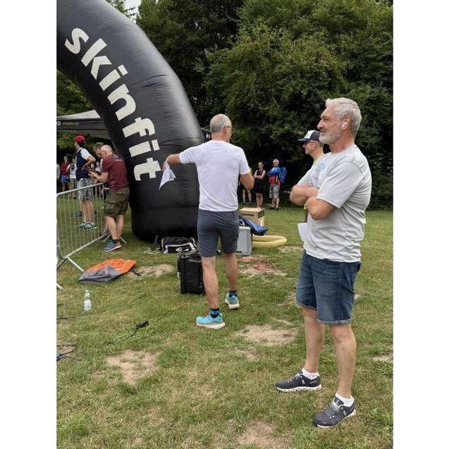 Zwei M&auml;nner stehen an einem Aufblasbogen mit der Aufschrift &bdquo;skinfit&ldquo; auf einer Rasenfl&auml;che, weitere Personen im Hintergrund.