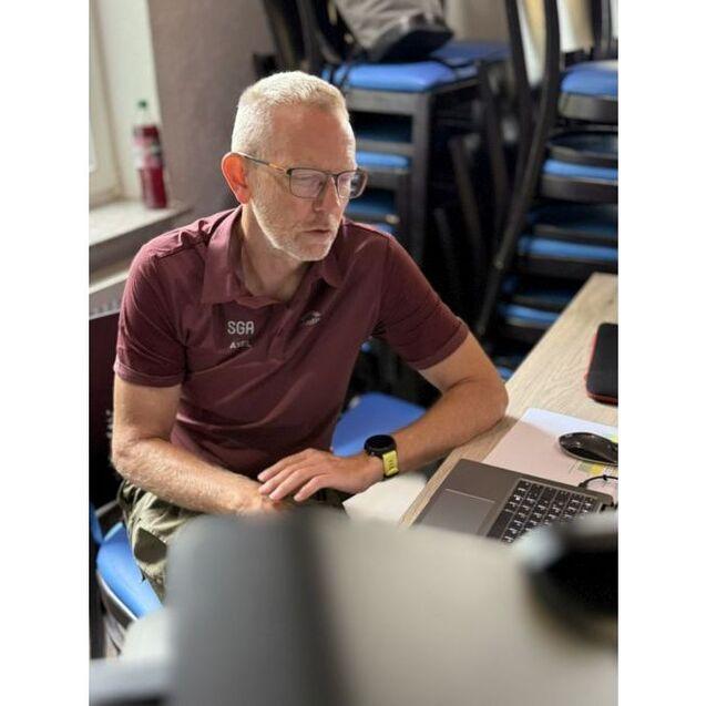 Mann mit Brille und Polohemd sitzt an einem Tisch und arbeitet am Laptop, im Hintergrund St&uuml;hle gestapelt.