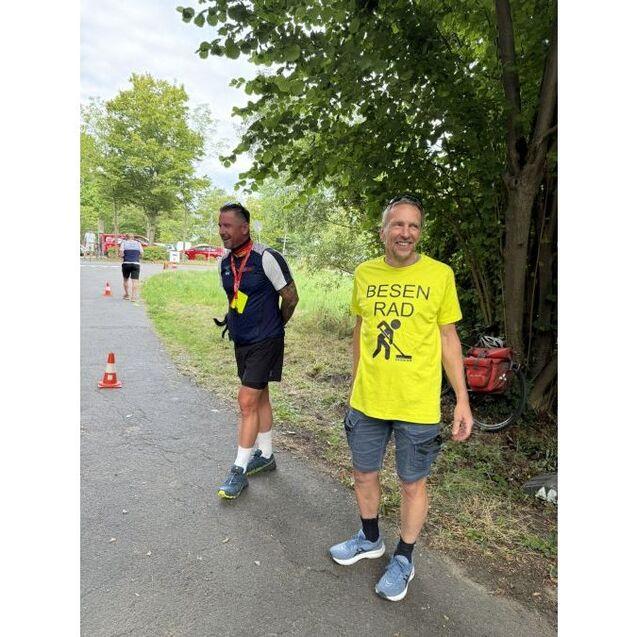 Zwei M&auml;nner stehen auf einem Weg. Einer tr&auml;gt ein leuchtend gelbes T-Shirt mit der Aufschrift &bdquo;BESEN RAD&ldquo;.