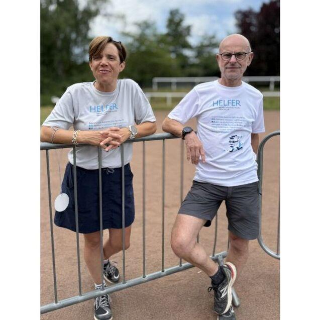 Zwei Personen in Sportbekleidung stehen an einem Zaun auf einem Sportplatz und l&auml;cheln in die Kamera.