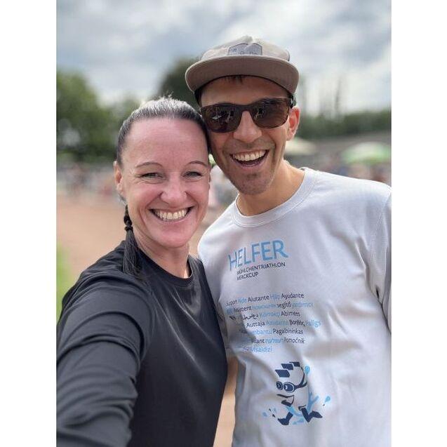 L&auml;chelndes Paar posiert f&uuml;r ein Selfie, w&auml;hrend im Hintergrund eine Veranstaltung im Freien stattfindet.