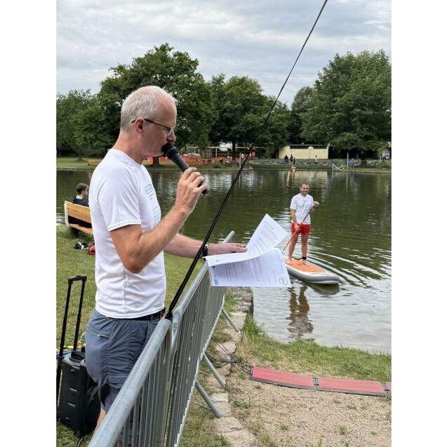 Mann mit Mikrofon steht vor einem See, w&auml;hrend ein Junge auf einem Board paddelt. B&auml;nke und B&auml;ume im Hintergrund sichtbar.