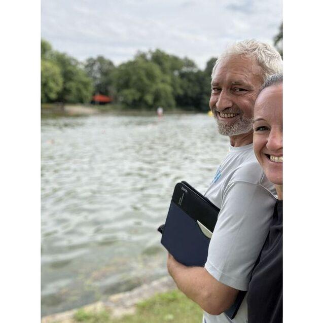 Zwei Personen stehen am Ufer eines Gew&auml;ssers und l&auml;cheln in die Kamera, mit einem Notizbuch in der Hand.