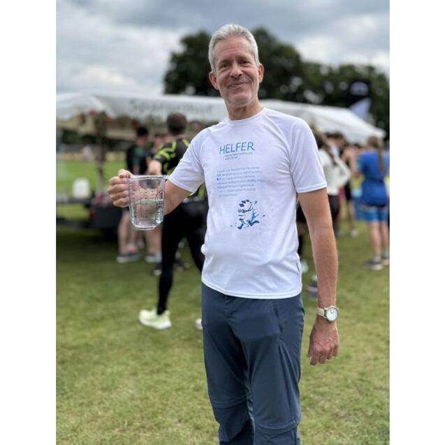 Mann in T-Shirt mit dem Wort &bdquo;HELFER&ldquo; h&auml;lt einen Messbecher mit Wasser auf einem Sportevent im Freien.