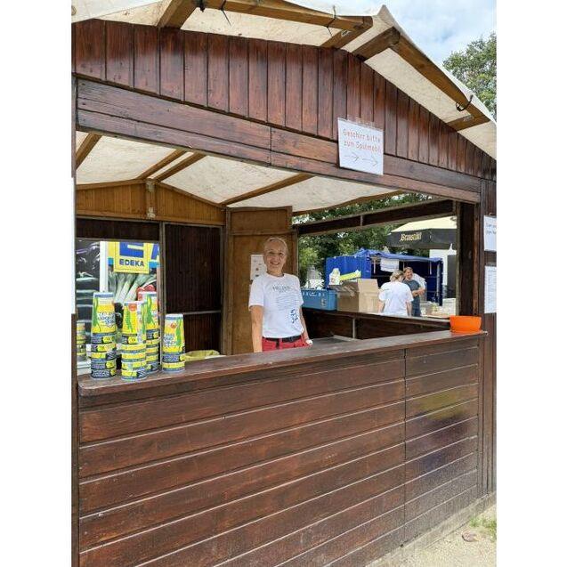 Ein Kassierer steht an einem Holzstand mit Dosen und einem EDEKA-Schild im Hintergrund.