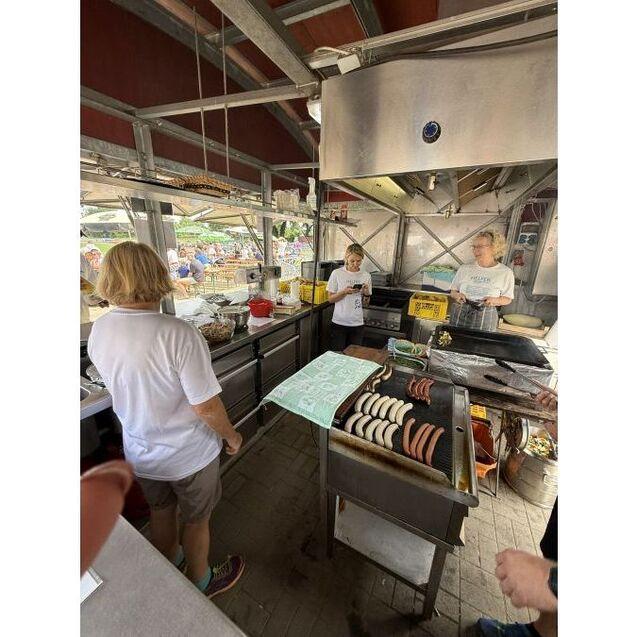 Drei Personen in wei&szlig;er Kleidung an einem Grillstand, Bratw&uuml;rste und Gem&uuml;se auf einem Grill, gesch&auml;ftige Marktatmosph&auml;re.