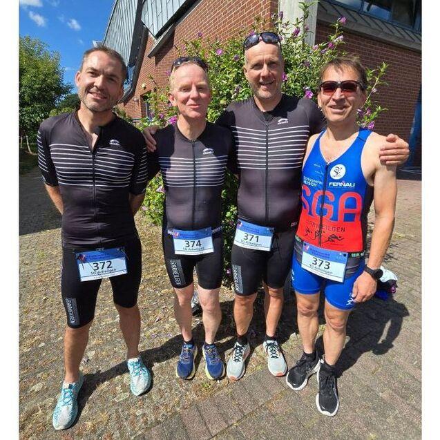 Vier M&auml;nner in Sportkleidung stehen l&auml;chelnd nebeneinander vor einer Wand mit Blumen. Jeder hat eine Startnummer auf der Brust.