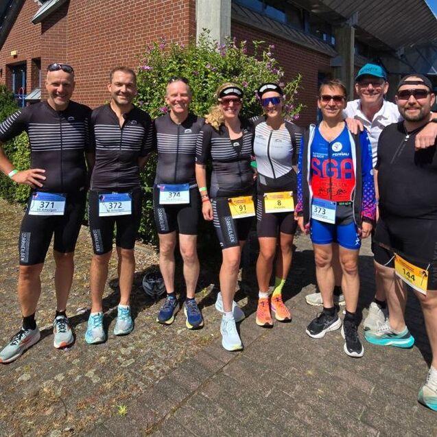 Gruppe von sieben Sportlern in Rennoutfits mit Startnummern vor einem Geb&auml;ude im Freien.