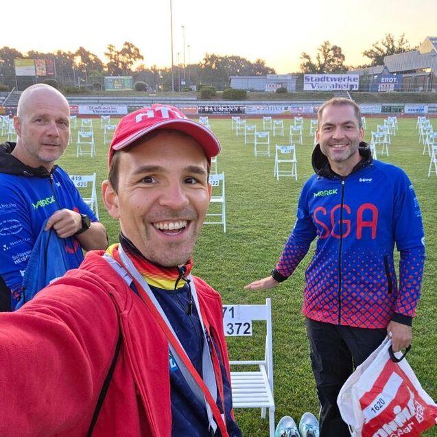 Drei M&auml;nner posieren im Stadion auf einer Wiese mit aufgestellten St&uuml;hlen, hinter ihnen beginnt die Morgensonne zu scheinen.