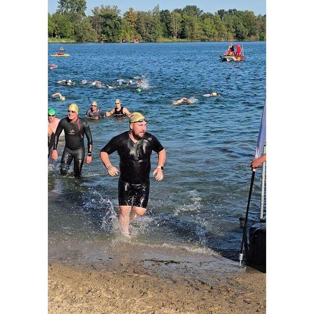 Mann im Neoprenanzug l&auml;uft aus einem See, w&auml;hrend weitere Schwimmer im Hintergrund Brustschwimmen.
