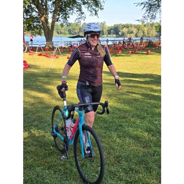 Frau in Sportbekleidung schiebt ein Rennrad auf einer Wiese am Ufer eines Sees. Hintergrund mit B&auml;umen und Fahrr&auml;dern.