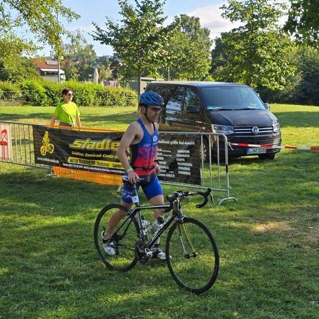 Mann in blauem Triathlonanzug schiebt ein Rennrad neben einer Werbebande in einem Park.