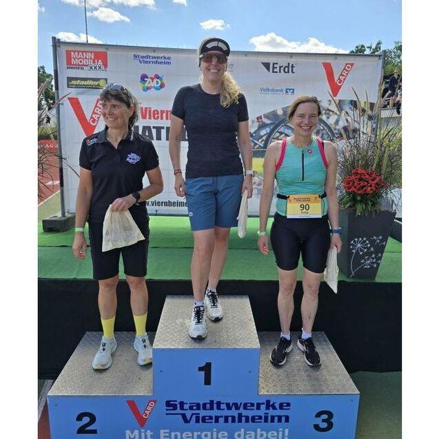 Drei Frauen stehen auf dem Siegerpodest bei einer Sportveranstaltung, jeder mit Urkunden in der Hand.