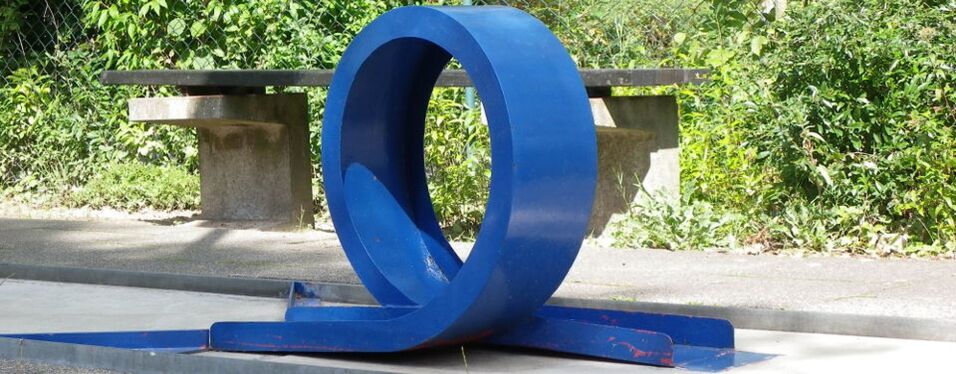 Blaue, skulpturale Sitzgelegenheit in Form eines Rings auf einem Gehweg, umgeben von Gr&uuml;npflanzen.