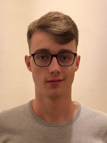 Portr&auml;t eines jungen Mannes mit Brille, kurzem Haar und grauem Oberteil vor einem hellen Hintergrund.