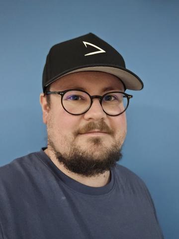 Portr&auml;t eines Mannes mit Brille und schwarzer M&uuml;tze vor einer blauen Wand. Er tr&auml;gt ein graues T-Shirt und l&auml;chelt.