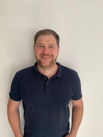 Mann mit kurzem, braunem Haar und Bart steht vor einer wei&szlig;en Wand, tr&auml;gt ein schwarzes Polohemd und l&auml;chelt.