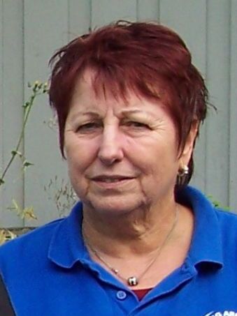 Portr&auml;t einer Frau mit kurzem, r&ouml;tlichem Haar, die ein blaues Oberteil tr&auml;gt, vor einer grauen Wand im Freien.