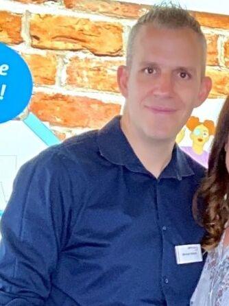 L&auml;chelnder Mann in blauem Hemd vor einer Backsteinwand, neben einer Frau und einem bunten Grafikhintergrund.