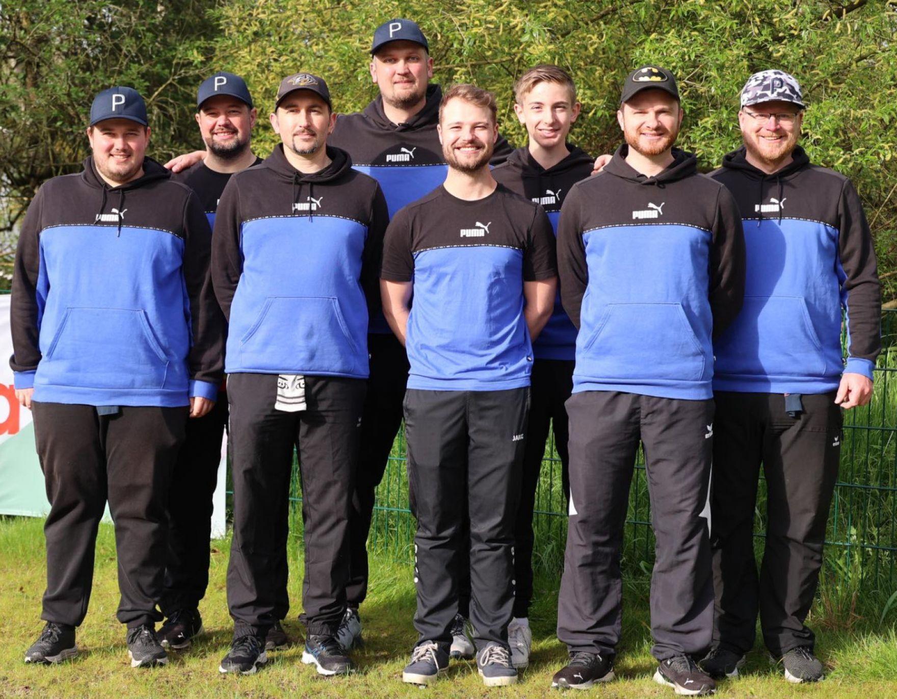 Gruppe von acht M&auml;nnern in einheitlicher Sportkleidung mit blauen und schwarzen Sweatshirts steht drau&szlig;en.