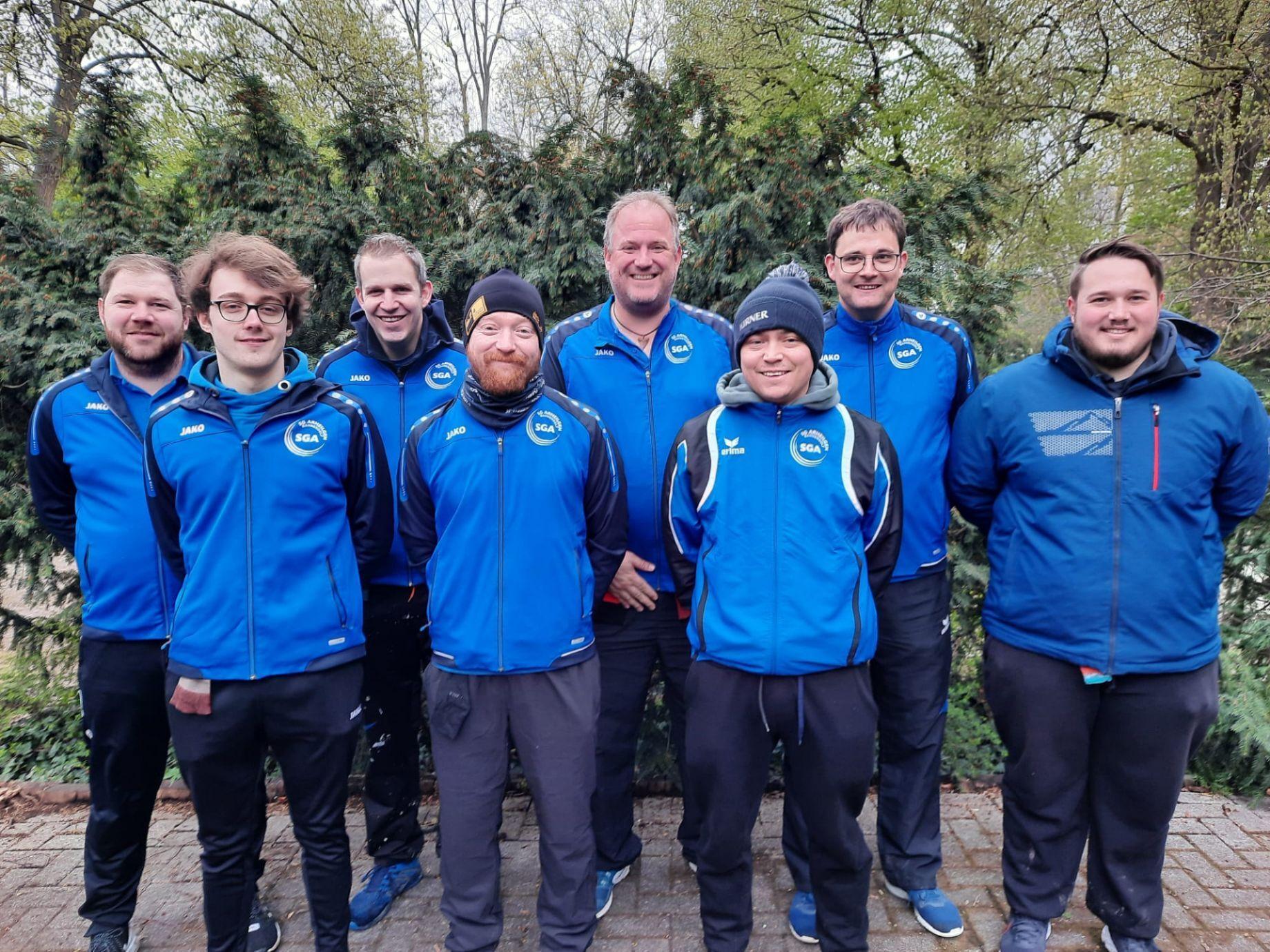 Gruppe von acht Personen in blauen Sportjacken vor einer gr&uuml;nen Hecke, einige l&auml;cheln in die Kamera.