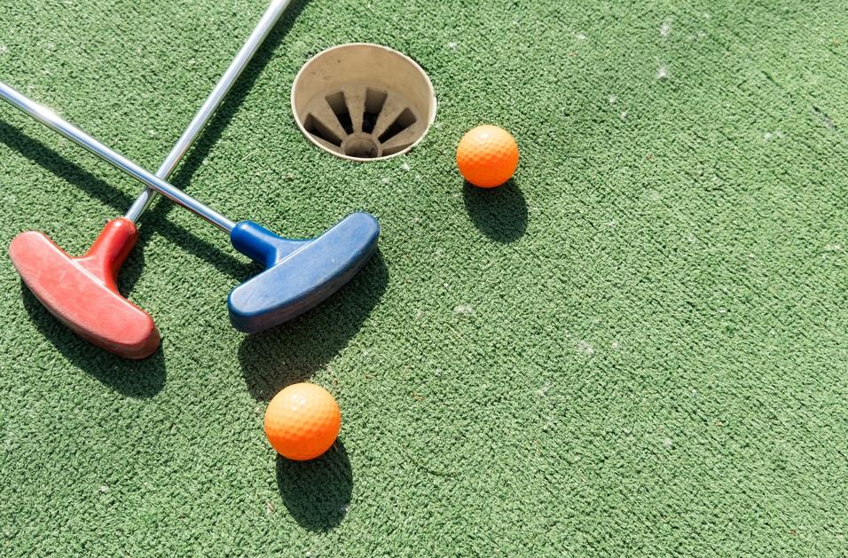 Zwei Putters, rot und blau, liegen neben einer orangefarbenen Golfball auf einem gr&uuml;nen Minigolfplatz.