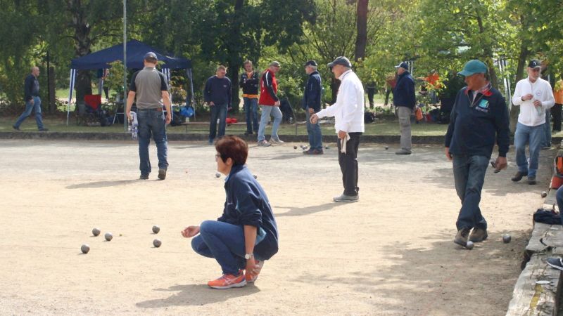 Mehrere Personen spielen Boule in einem Park, w&auml;hrend eine Frau konzentriert auf die Kugeln am Boden schaut.