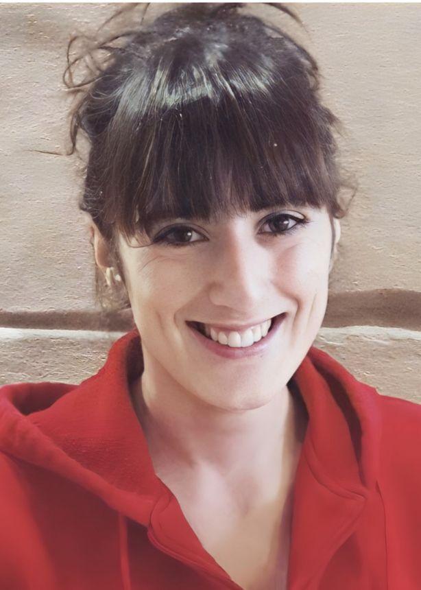 L&auml;chelnde Frau mit dunklen, lockigen Haaren, die ein rotes Sweatshirt tr&auml;gt, vor einer hellen Wand.