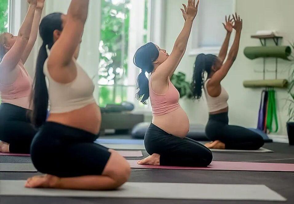 Schwangere Frauen praktizieren Yoga in einem hellen Raum mit gro&szlig;en Fenstern und Yogamatten auf dem Boden.