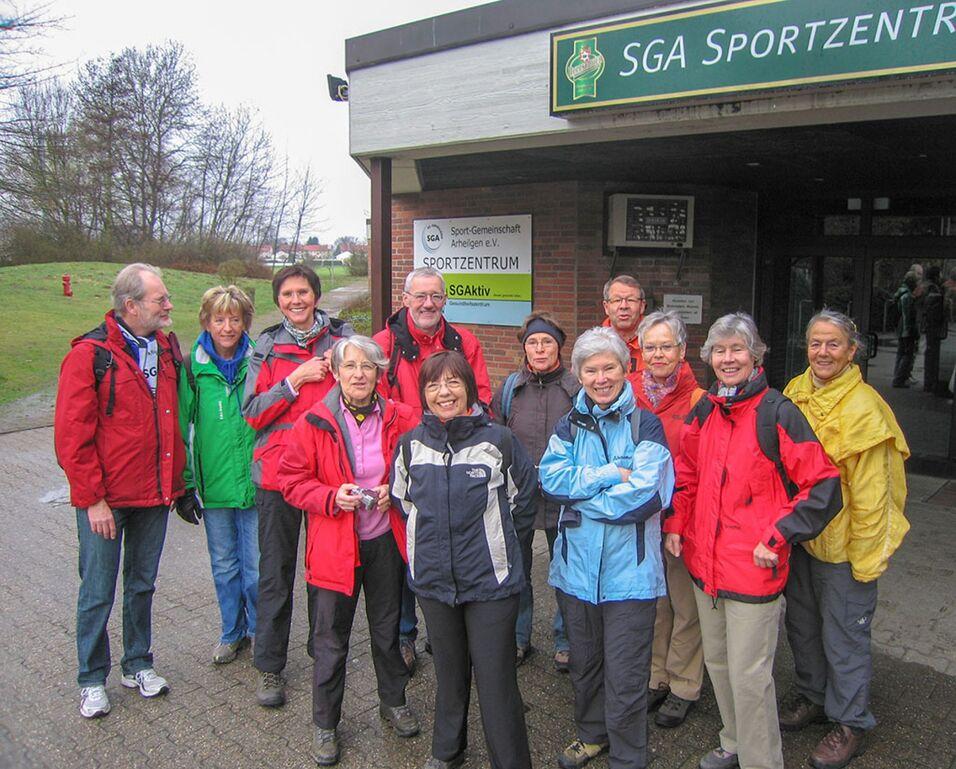 Gruppe von zw&ouml;lf Personen in Outdoor-Bekleidung vor dem SGA Sportzentrum, im Hintergrund gepflegte Landschaft.