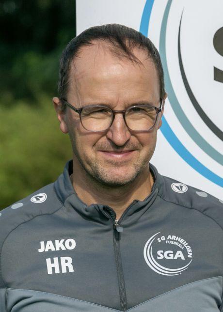 Portr&auml;t eines Mannes mit Brille, der ein graues Sportoberteil tr&auml;gt, vor einem sportlichen Hintergrund.