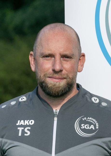 Portr&auml;t eines Mannes mit kurzem Bart, in Sportjacke der SGA, vor einem unifarbenen Hintergrund.