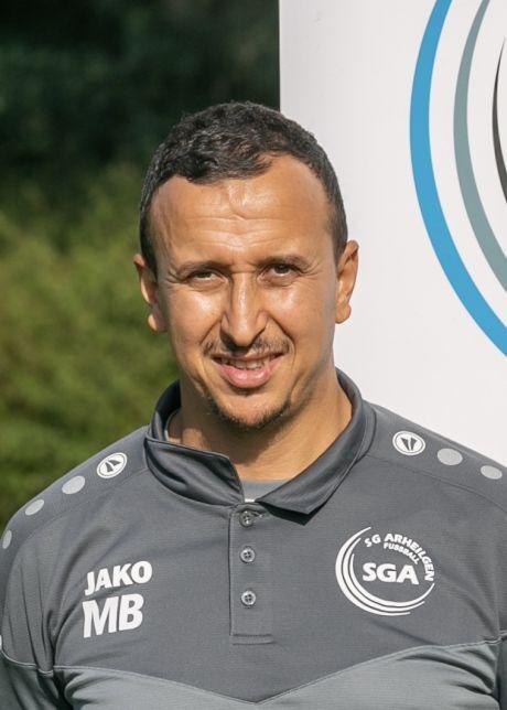 Portr&auml;t eines l&auml;chelnden Mannes in grauem Traineranzug vor einem Logo der SGA Arheilgen im Hintergrund.