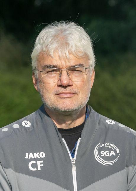 Portr&auml;t eines Mannes mit grauen Haaren, Brille und Trainingsanzug, vor unscharfem, gr&uuml;nem Hintergrund.