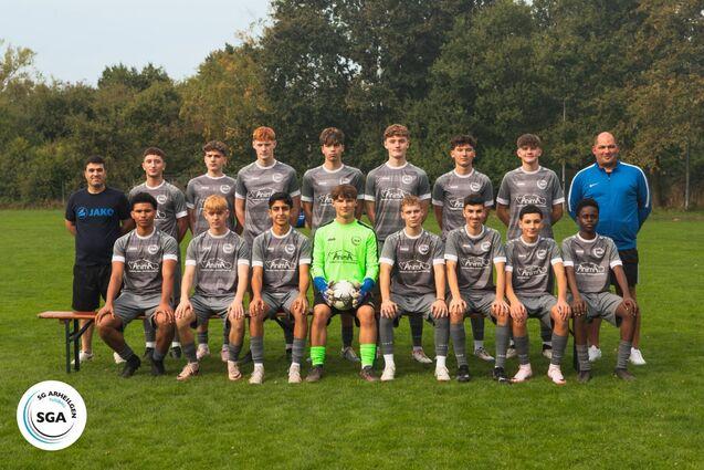Gruppenfoto einer Fu&szlig;ballmannschaft mit 13 Spielern und 2 Trainern auf einem Rasenfeld. Alle tragen graue Trikots.