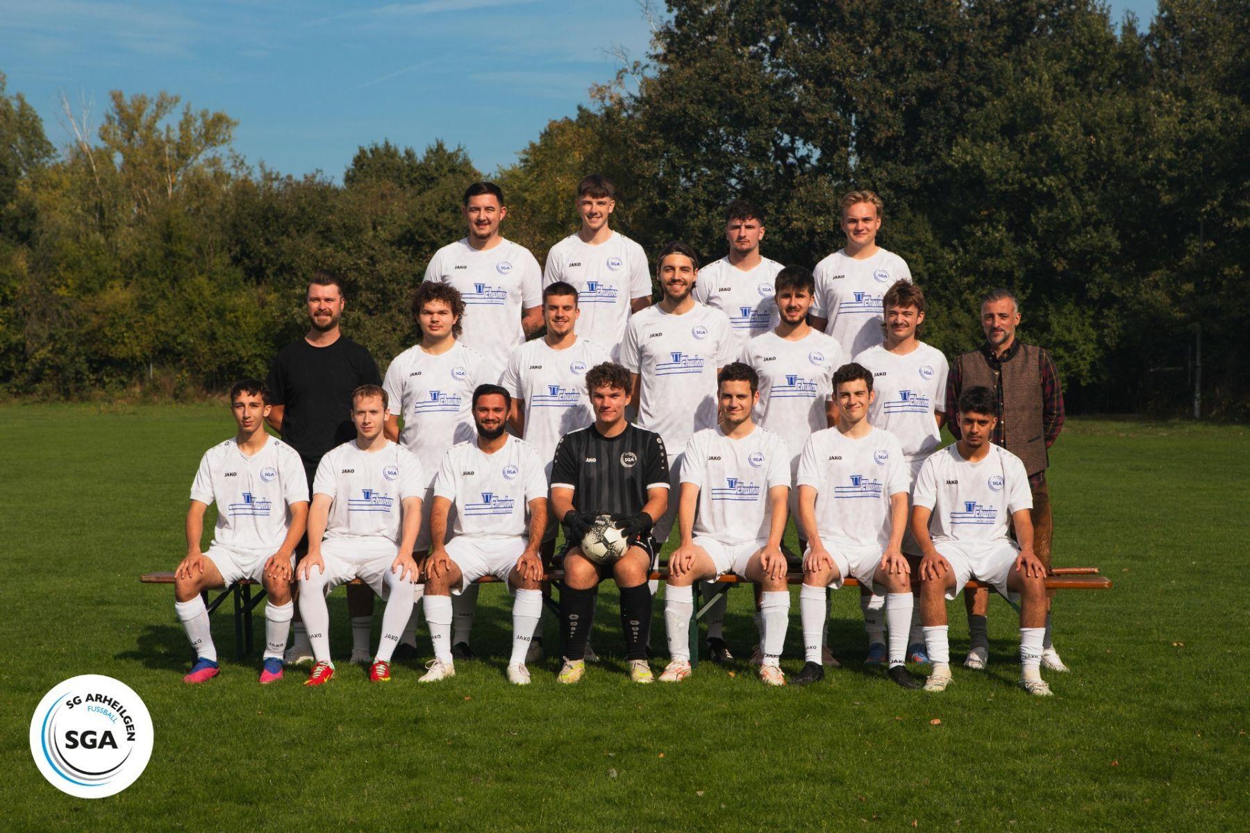 Mannschaftsfoto einer Fu&szlig;ballmannschaft in wei&szlig;en Trikots auf einer gr&uuml;nen Wiese, umgeben von B&auml;umen.