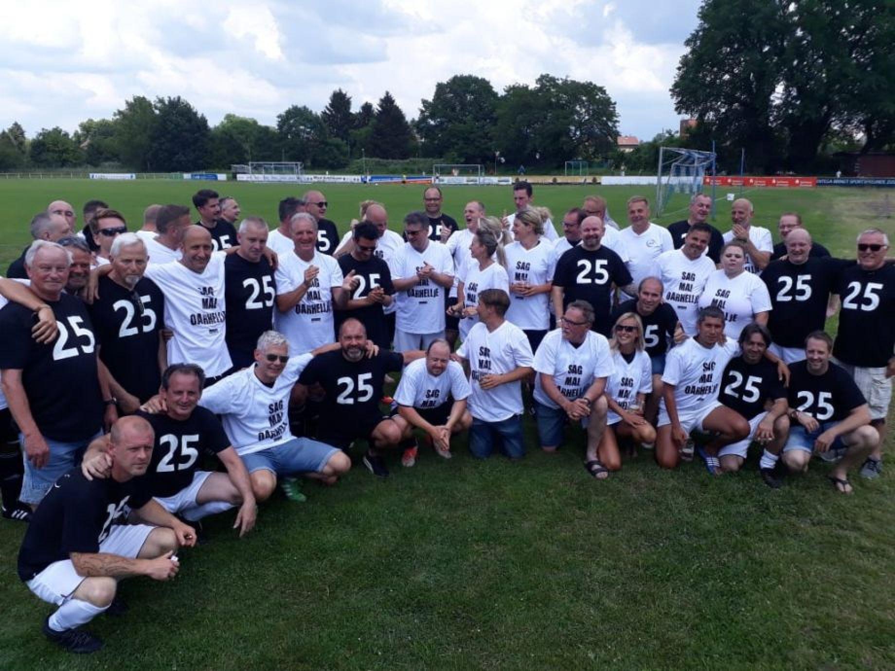 Gruppenfoto von M&auml;nnern und Frauen in T-Shirts mit der Zahl 25 auf einem gr&uuml;nen Fu&szlig;ballplatz.