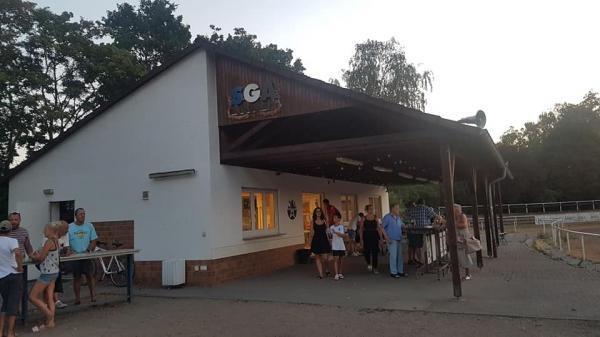 Menschen versammeln sich vor einem wei&szlig;en Geb&auml;ude mit &uuml;berdachter Terrasse, umgeben von B&auml;umen bei Sonnenuntergang.