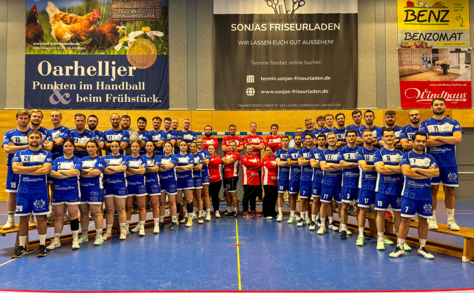 Mannschaftsfoto einer Handballmannschaft in blauen Trikots, posierend in einer Sporthalle mit Werbebannern im Hintergrund.