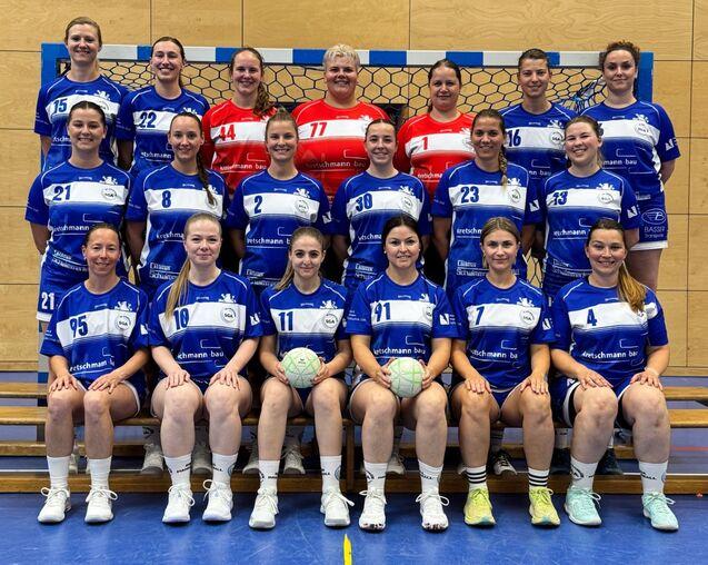 Gruppenfoto eines Handballteams in blauen und roten Trikots, l&auml;chelnd vor einem Handballtor in einer Sporthalle.