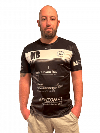 Mann mit Vollbart tr&auml;gt ein schwarz-wei&szlig;es T-Shirt mit Sponsorenlogos und steht vor einem einfarbigen Hintergrund.