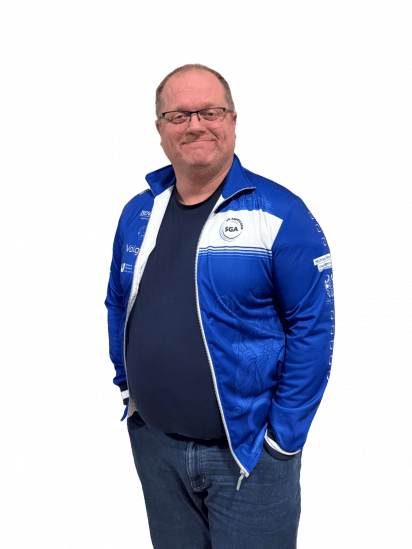 Mann in blauer Sportjacke mit Logo steht mit verschr&auml;nkten Armen und l&auml;chelt vor schwarzem Hintergrund.