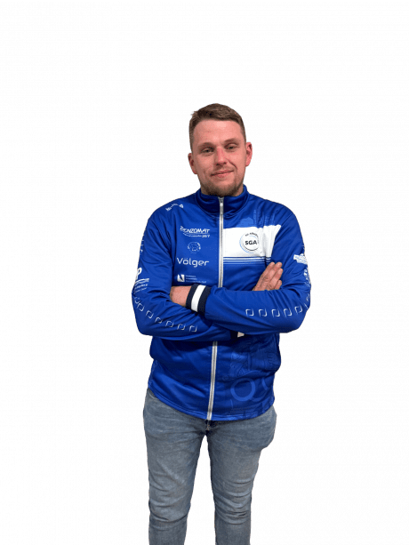 Junger Mann mit crossed arms in blauer Sportjacke, steht vor schwarzem Hintergrund.