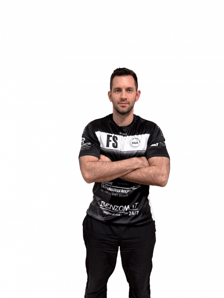 Mann mit kurzem Haar in Sporttrikot steht mit verschr&auml;nkten Armen vor neutralem Hintergrund.