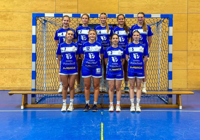 Handballmannschaft aus zehn Spielerinnen in blauen Trikots vor einem Tor in einer Sporthalle.