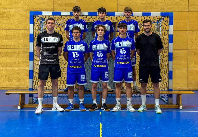 Mannschaftsfoto einer Handballmannschaft mit Trainer vor einem Tor in einer Sporthalle. Alle tragen blaue Trikots.
